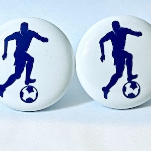 May include: Two white ceramic knobs with a blue silhouette of a soccer player dribbling the ball.