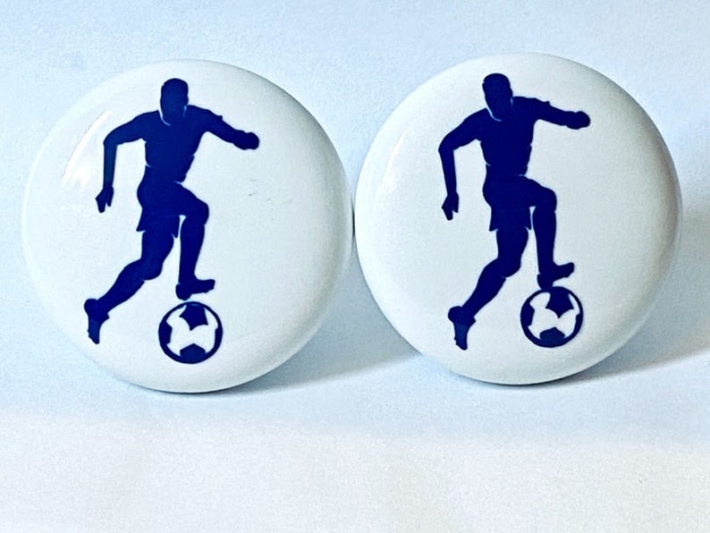 May include: Two white ceramic knobs with a blue silhouette of a soccer player dribbling the ball.