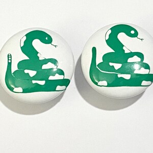 May include: Two white ceramic knobs with a green snake design. The snake is coiled and has white spots.