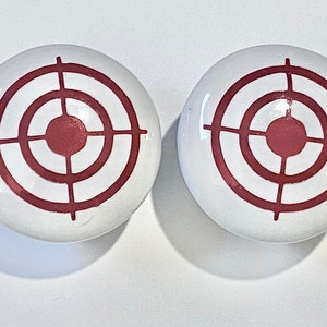May include: Two white ceramic knobs with a red target design. The target has three concentric circles and a crosshair in the center.