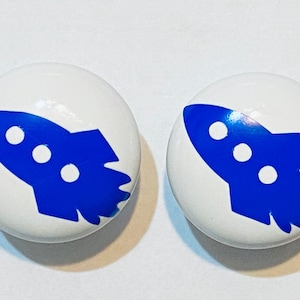 May include: Two white ceramic knobs with blue rocket ship designs. The rockets have three white circles on them.