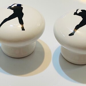Peut inclure: Deux boutons de cabinet en c&eacute;ramique blanche avec des silhouettes de joueurs de baseball noirs. Les joueurs de baseball sont en position de frappe.