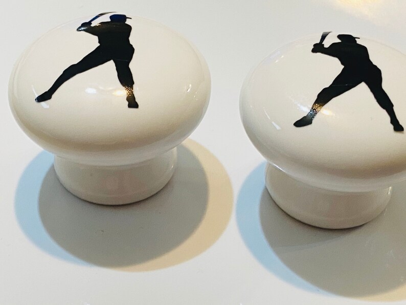 Peut inclure: Deux boutons de cabinet en c&eacute;ramique blanche avec des silhouettes de joueurs de baseball noirs. Les joueurs de baseball sont en position de frappe.