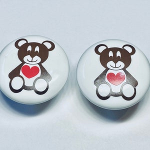 May include: Two white ceramic knobs with brown teddy bear designs holding red hearts. The teddy bears are smiling.
