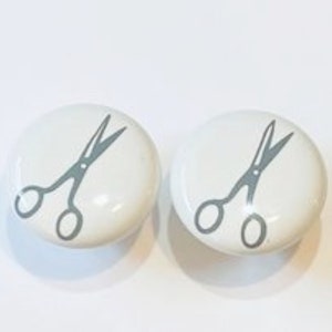 May include: Two white ceramic knobs with a gray silhouette of scissors on each.