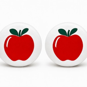 May include: Two round white ceramic knobs, each featuring a centered graphic of a bright red apple with two dark green leaves and a white highlight on the left side of the fruit, providing a clean and simple design for furniture hardware.