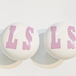 May include: Two white ceramic knobs with the letters "LS" in pink.