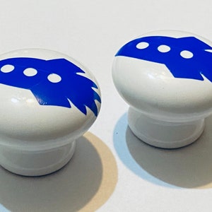Może przedstawiać: Dwa białe ceramiczne pokrętła z niebieskimi wzorami rakiet. Każde pokrętło ma trzy białe k&oacute;łka na rakiecie.