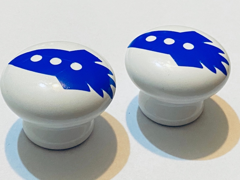 Może przedstawiać: Dwa białe ceramiczne pokrętła z niebieskimi wzorami rakiet. Każde pokrętło ma trzy białe k&oacute;łka na rakiecie.