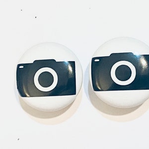 May include: Two white round buttons with a black camera icon on each. The camera icon has a white circle in the center.