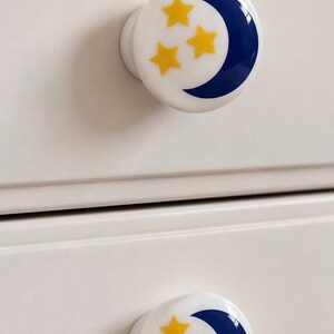 May include: Two round white ceramic drawer knobs, each featuring a glossy finish with a dark blue crescent moon graphic and three small yellow five-pointed stars printed on the front surface, attached to a light-colored wooden furniture drawer.