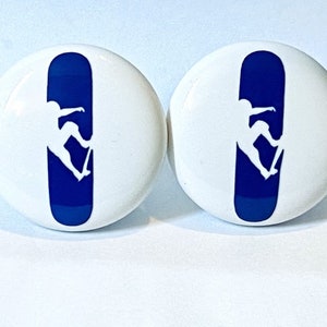 May include: Two white ceramic round buttons with a blue skateboarder silhouette on a blue background.