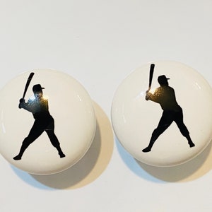 May include: Two white ceramic knobs with black silhouettes of baseball players holding bats.