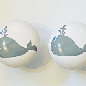 May include: Two white ceramic drawer knobs with a gray whale design. The whale has a small, black eye and a spout on top of its head.