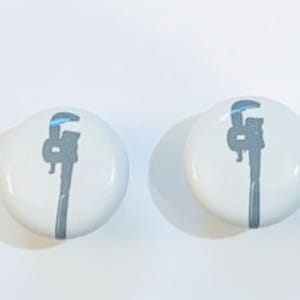 May include: Two white round ceramic knobs with a gray wrench design.