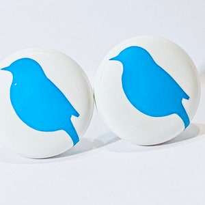 May include: Two white round buttons with a blue bird silhouette on each. The birds are facing the right side of the image.
