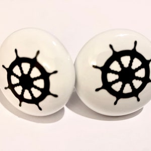 May include: Two white ceramic knobs with a black ship's wheel design.