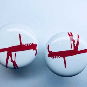 May include: Two white, round cabinet knobs with a red graphic design. The design appears to be a stylized animal or creature. The knobs are likely used for furniture or decorative purposes.