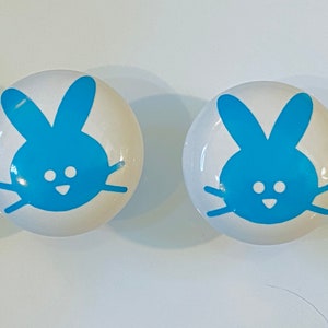 May include: Two white ceramic knobs with blue bunny designs. The bunnies have white eyes and a small mouth.