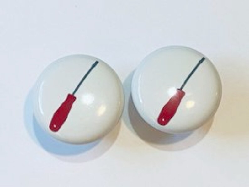 May include: Two white ceramic knobs with a red screwdriver design. The screwdriver is outlined in black and has a blue handle.