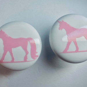 May include: Two white ceramic knobs with pink unicorn silhouettes. The unicorns are facing the right side of the knobs.