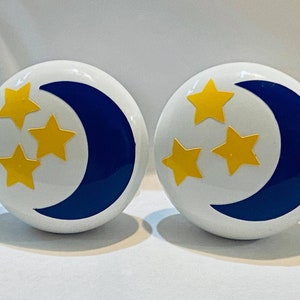 May include: Two white ceramic knobs with a blue crescent moon and five yellow stars. The knobs are round and have a smooth surface.