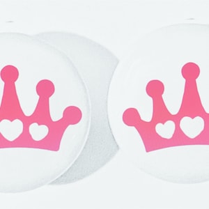 May include: Two white round stickers with a pink crown design. The crown has three white hearts in the center.