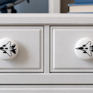 May include: Two round white ceramic cabinet knobs, each featuring a centered black graphic illustration of a fighter jet. The knobs are mounted onto a light gray wooden drawer front with a rectangular panel design and a smooth matte finish.