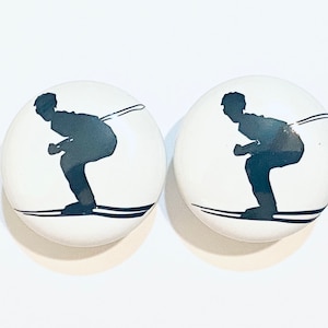 May include: Two white ceramic knobs with a black silhouette of a skier on each. The skier is in a crouched position with skis and ski poles.