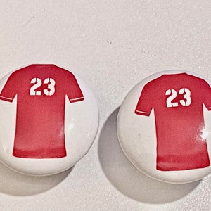 May include: Two white round buttons with red and white jersey designs. The jerseys have the number 23 printed on the front.