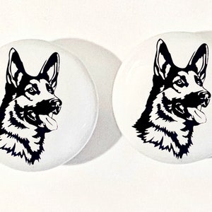 May include: Two white ceramic coasters with a black silhouette of a German Shepherd dog's head. The dog is looking to the right and has its tongue sticking out.