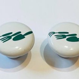 May include: Two white ceramic knobs with a green silhouette of gardening tools, including a rake, a fork, and a trowel.
