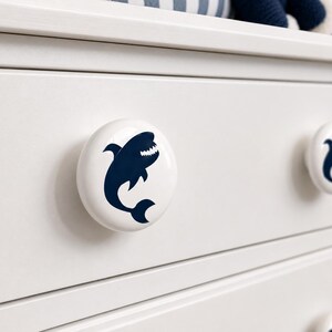 May include: A white circular cabinet knob featuring a dark navy blue graphic of a shark with an open mouth showing teeth. The knob is mounted on a white drawer front, with a second identical shark knob visible to the right on the same drawer.