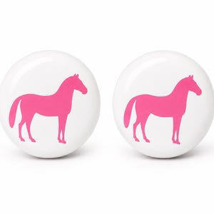 May include: Two circular white buttons, each featuring a solid, vibrant pink silhouette of a horse standing in profile. The buttons have a smooth, glossy finish and a simple, clean design, showcasing the bright pink equine graphic clearly.