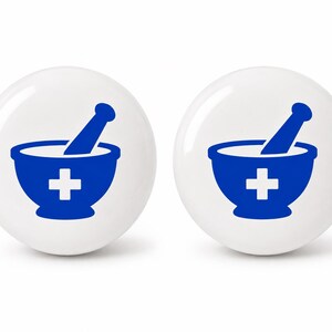 May include: Two round, white ceramic cabinet knobs, each featuring a blue graphic of a mortar and pestle with a white cross symbol centered on the front. The design is printed clearly on the smooth, glossy surface of these spherical hardware pieces.