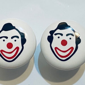 May include: Two white ceramic knobs with a black and red clown face design. The clown has a black mustache and eyebrows, a red nose, and a red smile.