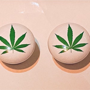 May include: Two white ceramic knobs with a green marijuana leaf design.