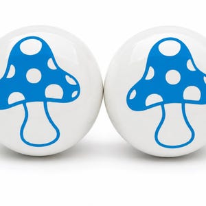 May include: Two round white ceramic knobs, each featuring a bright blue mushroom graphic with white polka dots on the cap. The knobs have a smooth, glossy finish and a spherical shape, providing a clean and simple design for furniture hardware.