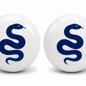 May include: Two spherical white ceramic knobs, each featuring a dark blue graphic of a coiled snake. The smooth, glossy surface of the round knobs provides a clean contrast to the bold, centered serpent illustration, creating a uniform set.