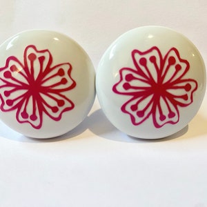 May include: Two white ceramic knobs with a pink floral design. The knobs are round and have a smooth surface.