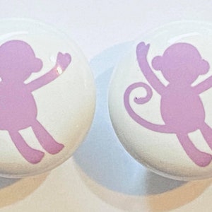 May include: Two white ceramic knobs with a pink monkey silhouette design. The monkeys are facing opposite directions and have their arms raised.