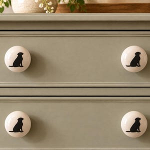 May include: Four round, off-white ceramic cabinet knobs are attached to a sage green dresser with two drawers. Each knob features a black silhouette of a sitting dog, providing a consistent decorative detail across the furniture piece.