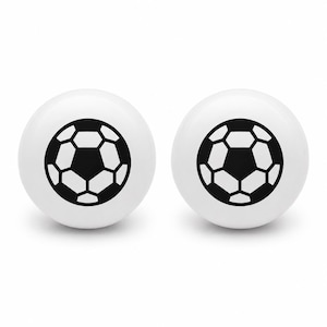 May include: Two round white cabinet knobs, each featuring a centered black and white graphic print of a soccer ball pattern. The knobs have a smooth, glossy finish and a spherical shape, designed for attachment to furniture or cabinetry doors.
