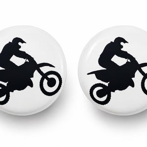 May include: Two circular white pin-back buttons, each featuring a black silhouette graphic of a person riding a motocross dirt bike. The design shows the rider in a seated position on the motorcycle with wheels clearly visible in profile view.
