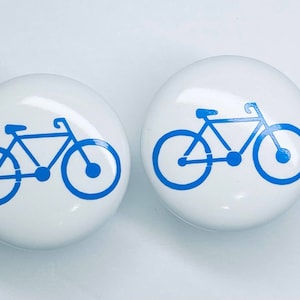 May include: Two white ceramic knobs with a blue bicycle design. The bicycle is a simple silhouette with two wheels, handlebars, and a seat.