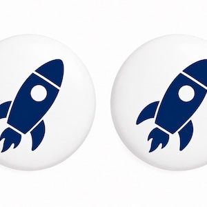 May include: Two circular white buttons, each featuring a dark blue graphic of a rocket ship. The rocket design includes a pointed nose cone, a circular window, side fins, and flames exiting the base, centered on the white circular background.