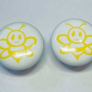 May include: Two white ceramic knobs with a yellow bee design. The bees have a smiling face and black stripes.
