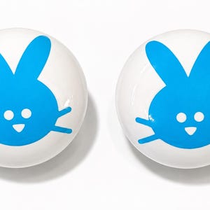 May include: Two round, white ceramic knobs, each featuring a bright blue graphic of a rabbit face. The rabbit design includes two tall ears, two small circular eyes, and a triangular nose, all rendered in a solid, vibrant blue color.