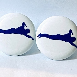 May include: Two white ceramic round buttons with a blue silhouette of a person swimming in a freestyle stroke.