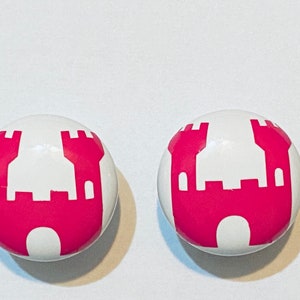 May include: Two white, round cabinet knobs with a magenta castle design. The castle illustration is centered on each knob, featuring a detailed silhouette of turrets and a central gate. These decorative knobs are suitable for furniture.